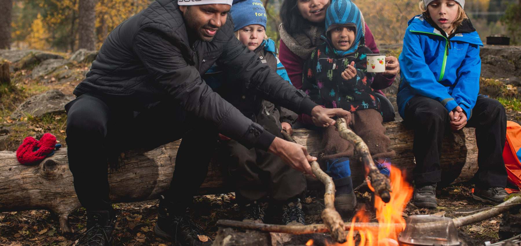 Two adults and three kids preparing food over fire, at a campsite.