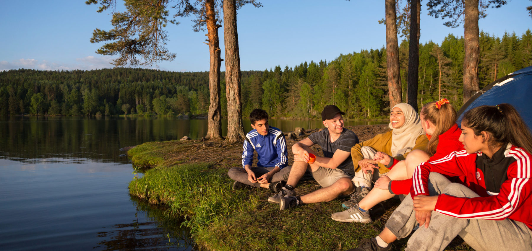 Ungdommer sitter foran telt ved et skogvann.