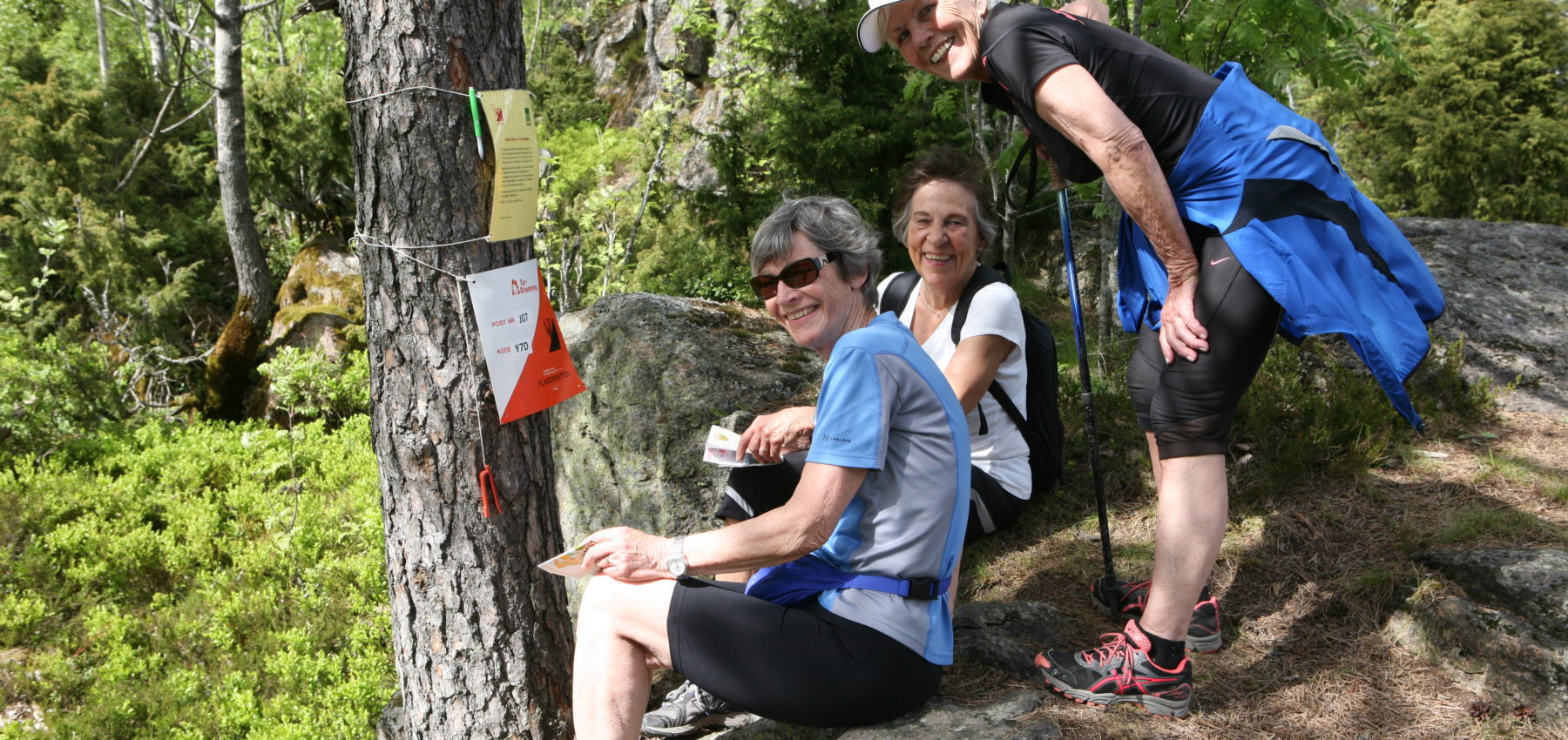 Tre damer på turorientering i skogen, smiler og ser inn i kamera.