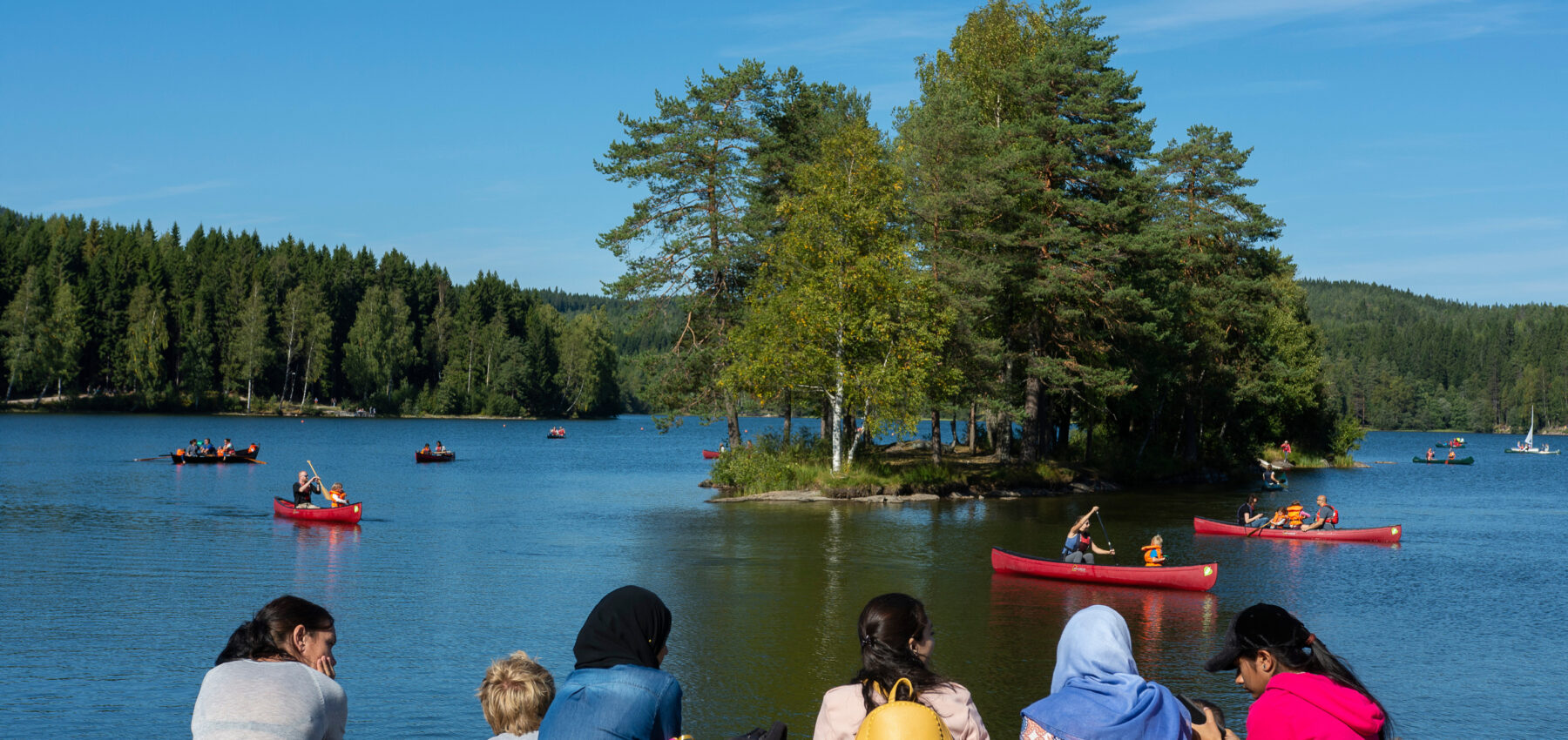 En gruppe sitter ved vannkanten og ser på barn som padler i kano på Sognsvann.